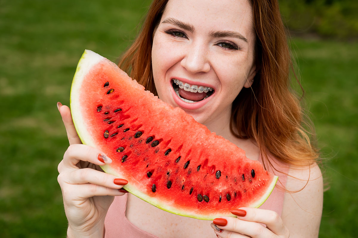 Close-up portrait of red-haired young woman with braces eating watermelon outdoors.