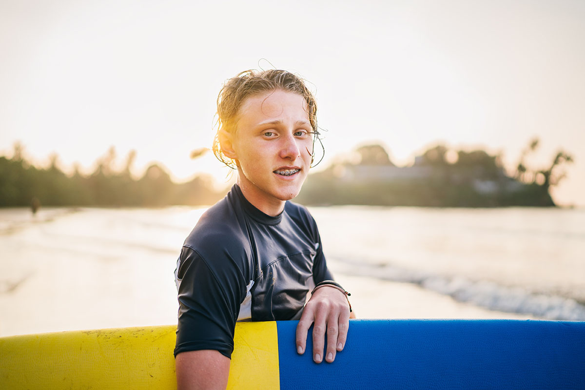 Teen boy with dental braces holding his surfboard.