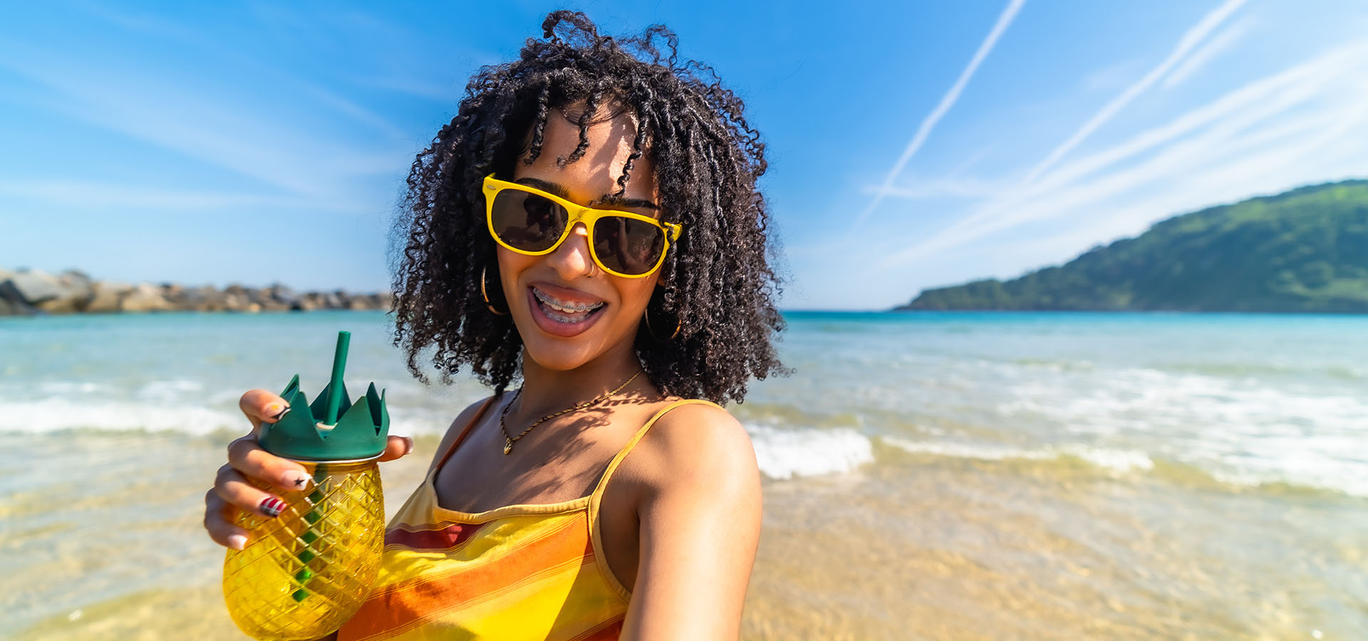 Young woman wearing sunglasses and braces, holding a pineapple shaped drink, smiling brightly at the camera while standing in shallow water at a sunny tropical beach
